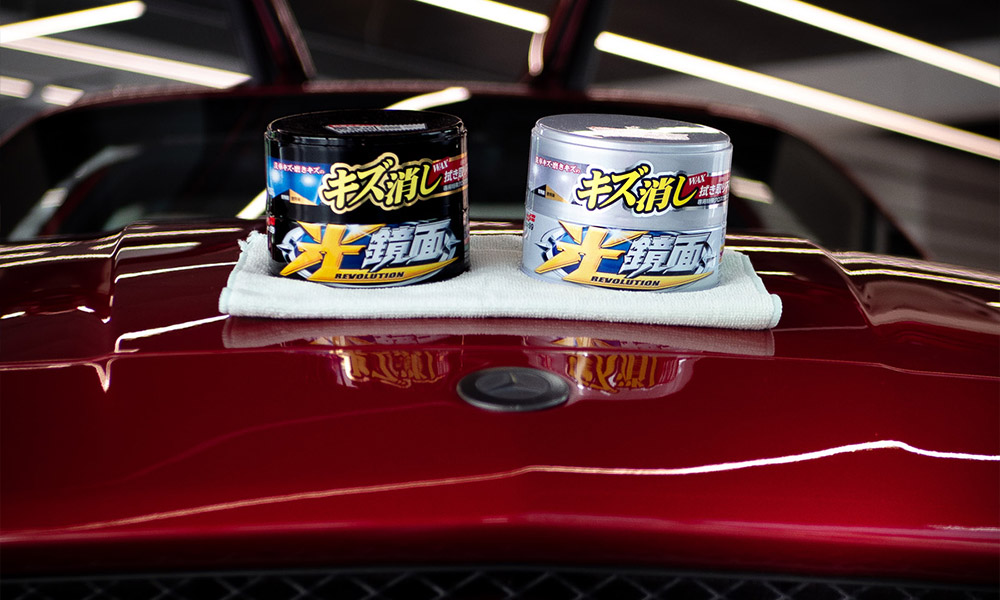 Photo of Soft99 car waxes Scratch Clear (variants for dark and light paintworks) placed on a microfibre on the bonnet of a shiny sport car.